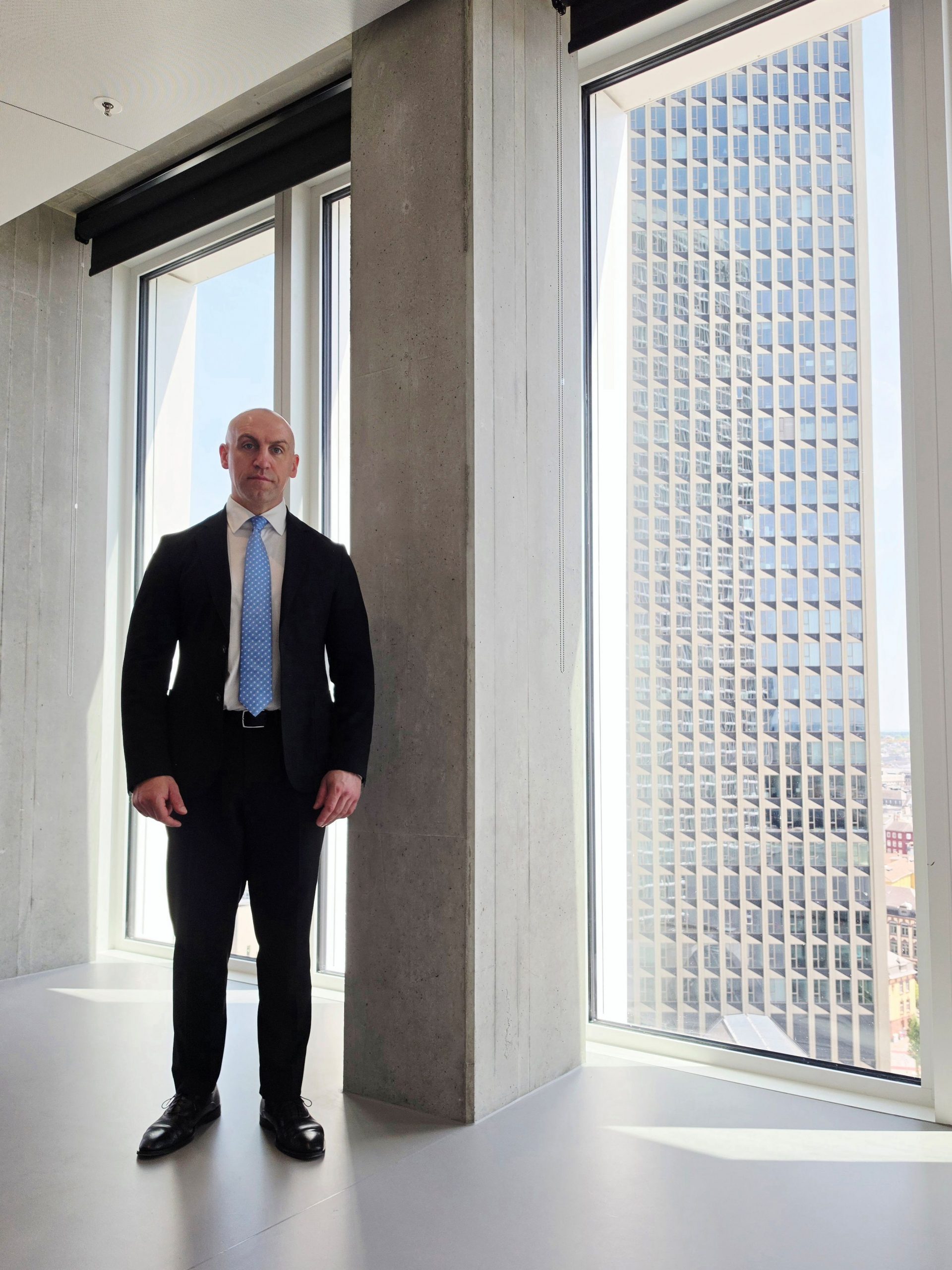 Ronny Kazyska in einem repräsentativen Hochhausinnenraum mit Blick auf Frankfurter Hochhausarchitektur zum Grundsatz Finanzierbarkeit bei Investmentimmobilien.