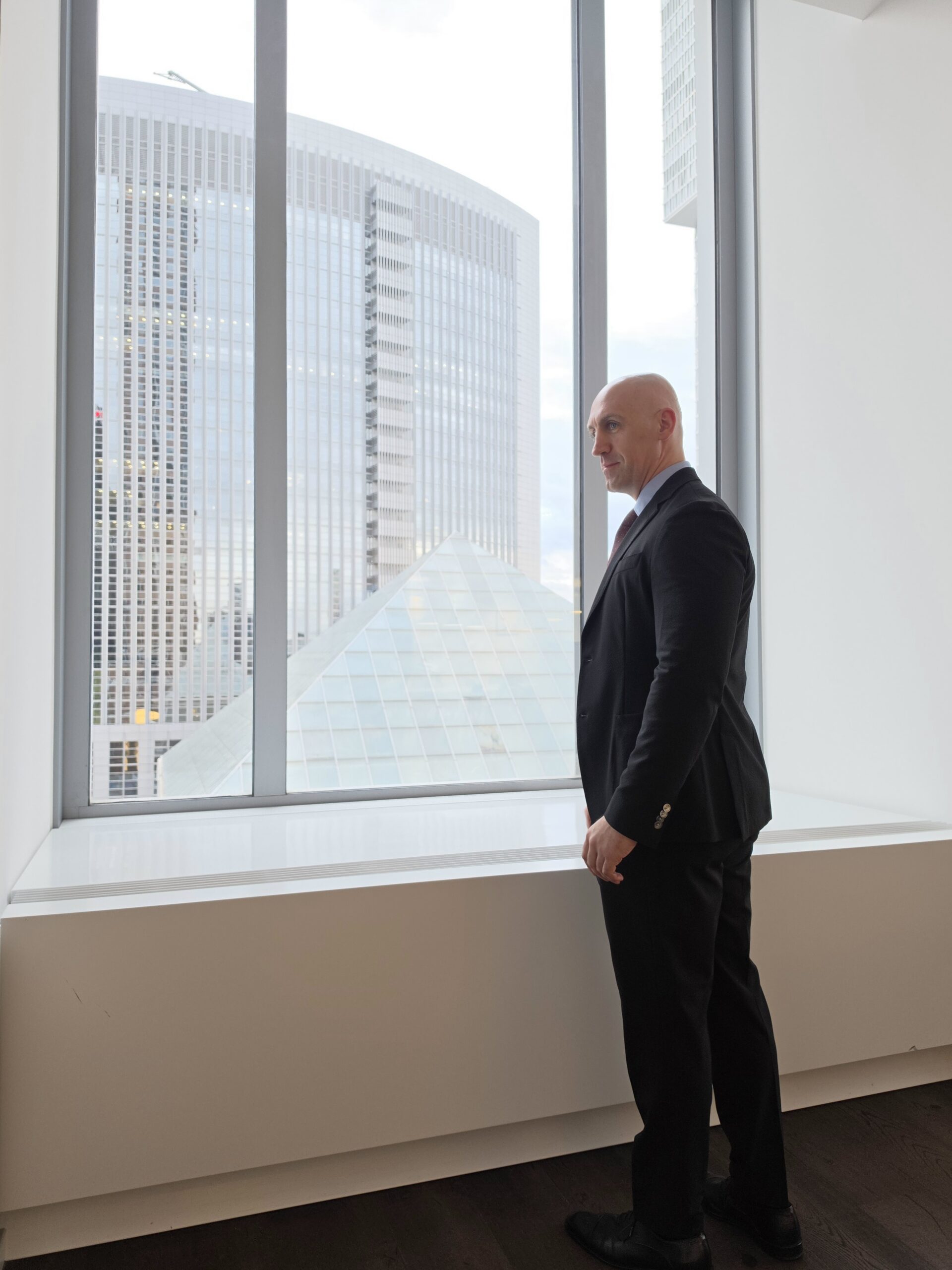 Ronny Kazyska am Fenster eines Büros im Frankfurter Messeturm mit Blick auf Hochhäuser des Frankfurter Bankenviertels.